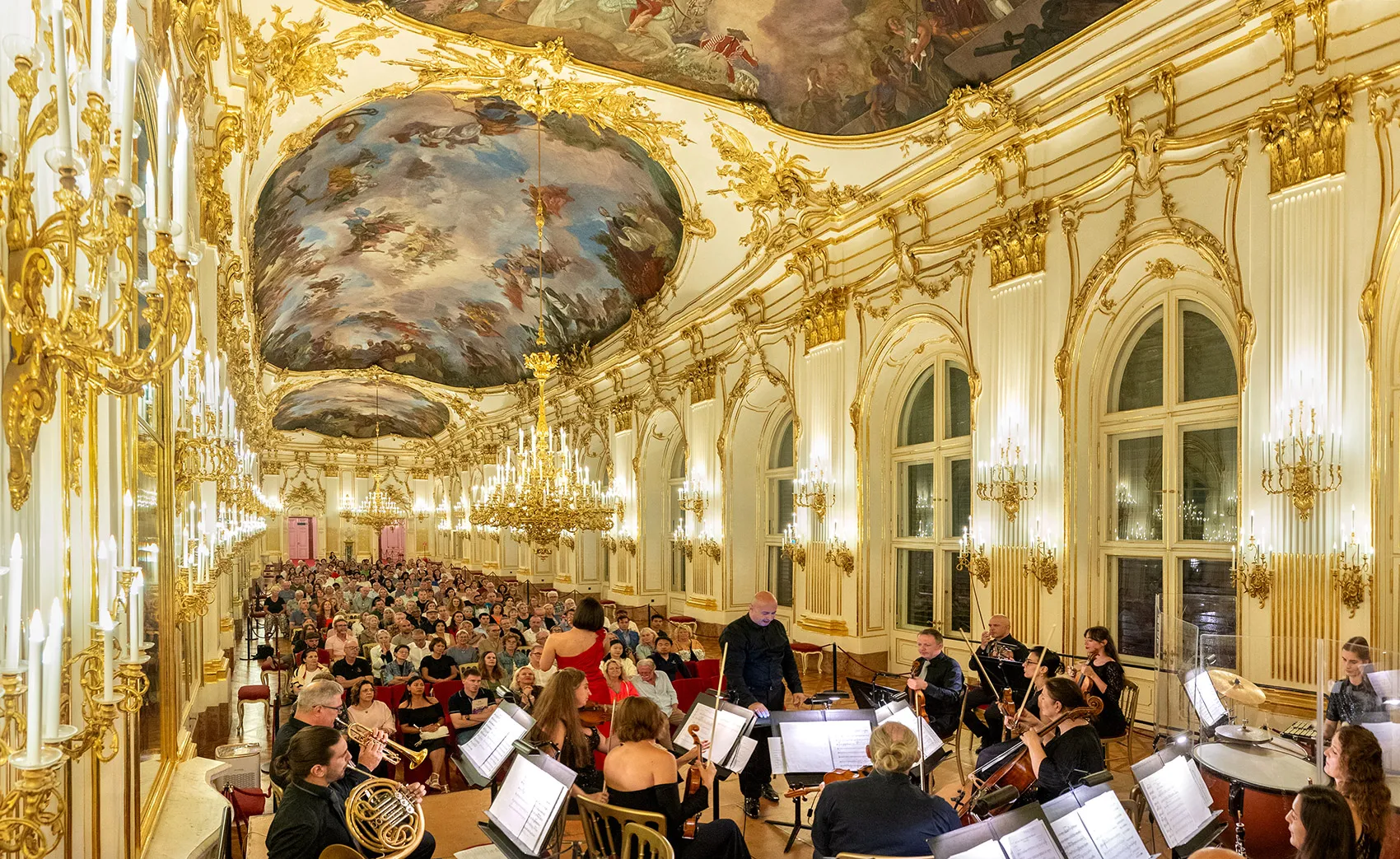 The Great Gallery of Schönbrunn during a concert: the white-and-gold Rococo hall under Guglielmi’s ceiling fresco, the orchestra and the audience beneath it.